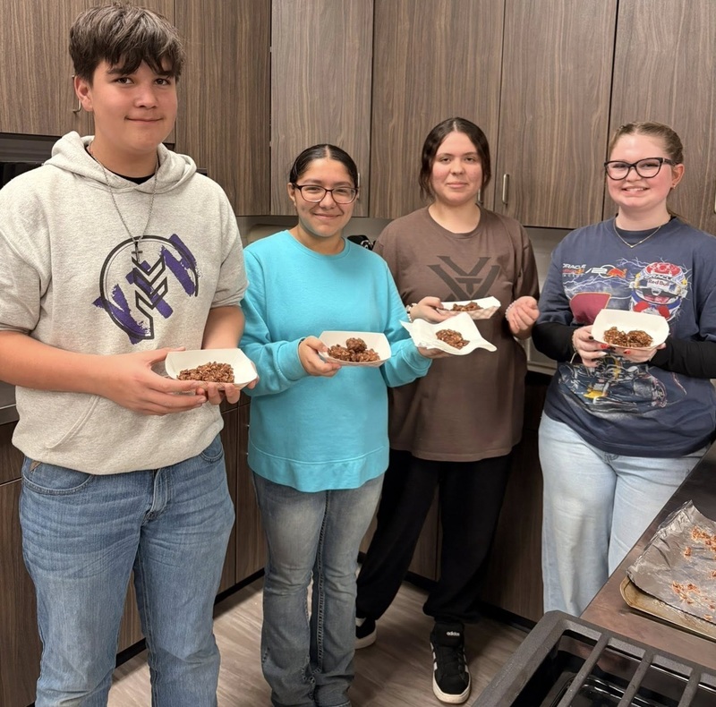 One teen boy and three teen girls smile for the camera in a kitchen classroom. he students hold paper trays containing chocolate cookies.