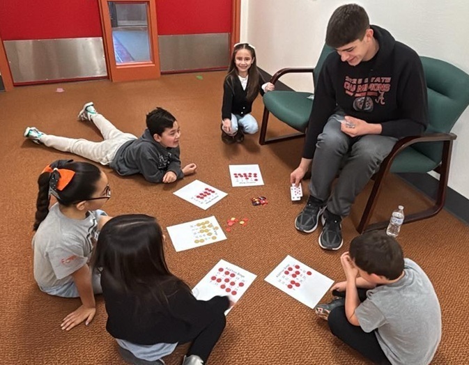 Two young male and three young female students sit on the floor in a semicircle around a teen boy seated on a chair. The students have worksheets in front of them.