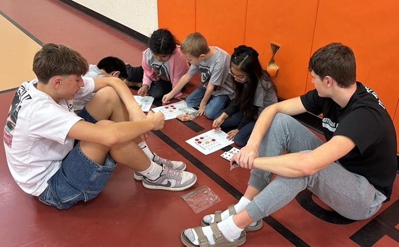 One young male and two young female students sit on the floor with their attention on worksheets as two teen boys also seated on the floor look on.