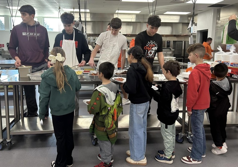 A line of young children files past a long table from which four teen boys fill the children's plates with breakfast foods.