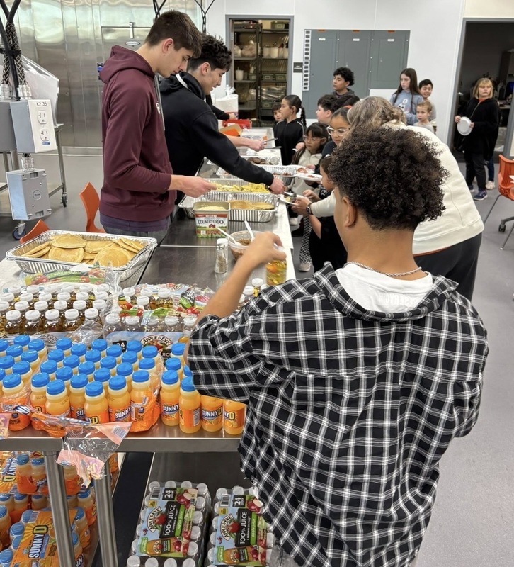 A teen boy, seen from behind, arranges small orange juice bottles on a table. In the background, other teen boys can be seen serving food to young students.