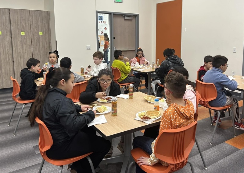 Young students are seen seated in orange chairs at multiple tables in a large room. The children are eating breakfast foods.