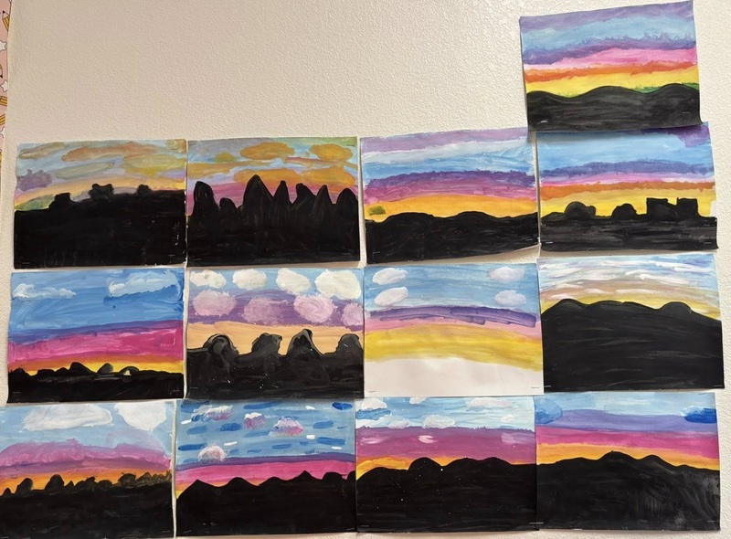 Multiple children's drawings hanging on a wall depict colorful sunsets against silhouetted desert landscapes.