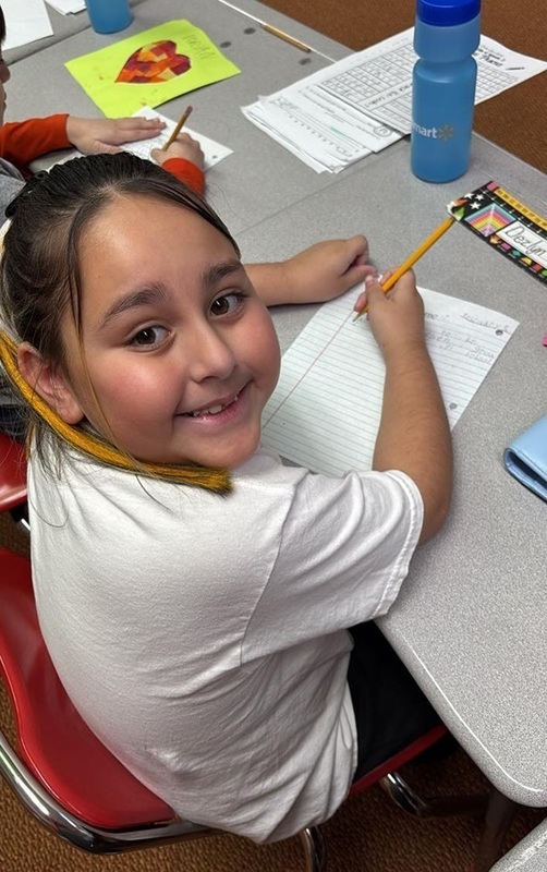 A young girl seated at a desk turns and smiles for the camera as she writes on a piece of paper with a pencil.