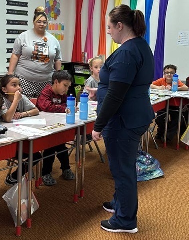 A woman in dark-blue scrubs stands in front of a row of desks and speaks to a young girl, who is listening attentively. 