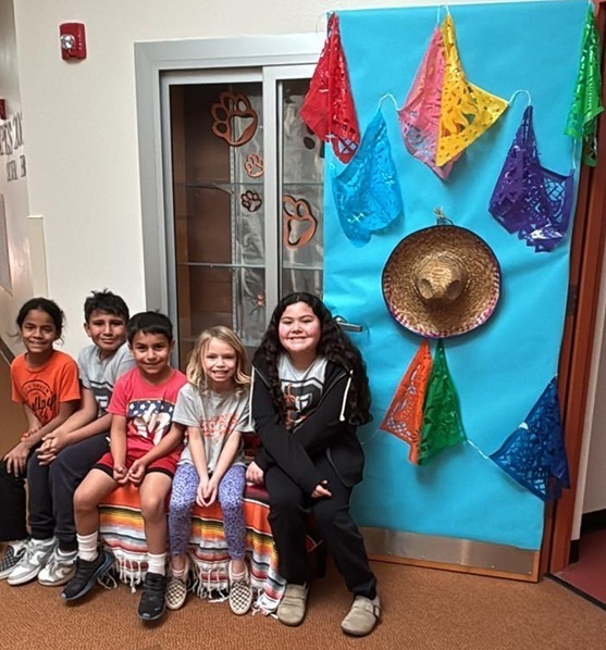 Two male and three female students sit on a bench and smile for the camera alongside a door decorated in brightly colored papel picado and a sombrero.