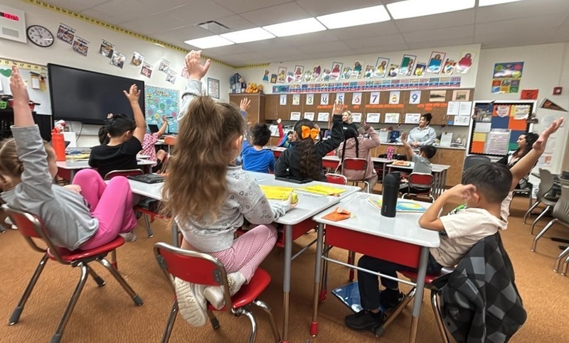 A classroom of young students, seen from behind, raise their hands to ask questions of two women at the front of the room.