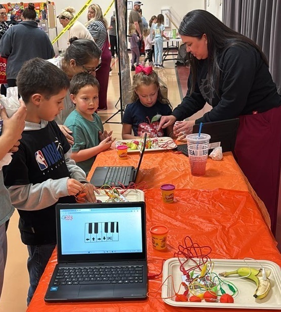 Children gather around a table manned by a female teacher. On the table sit two laptops and piece of fruit with wires protruding from them.