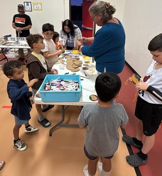 Several young children gather around a table manned by a female teacher. On the table sit several cardboard circles, yellow string, and a tub full of colored markers.