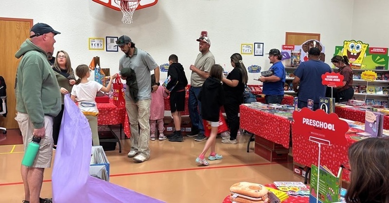 Children and adults mill around a school gym filled with tables. On the tables sit books.