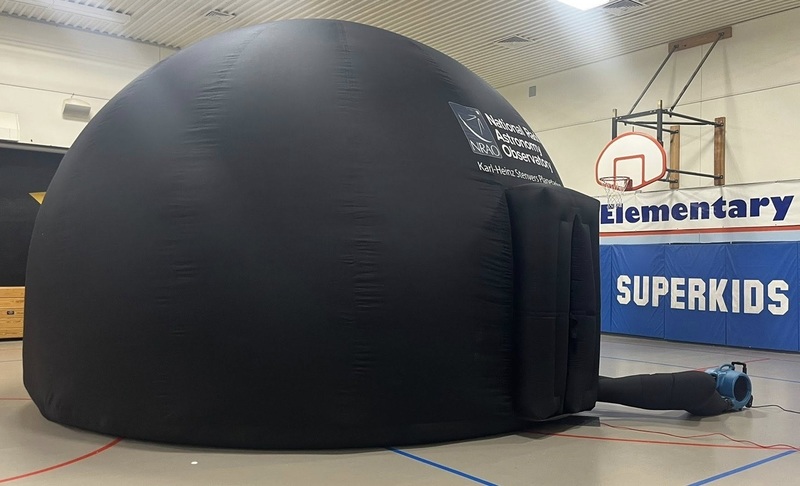 A large black inflatable dome is pictured in a school gymnasium.