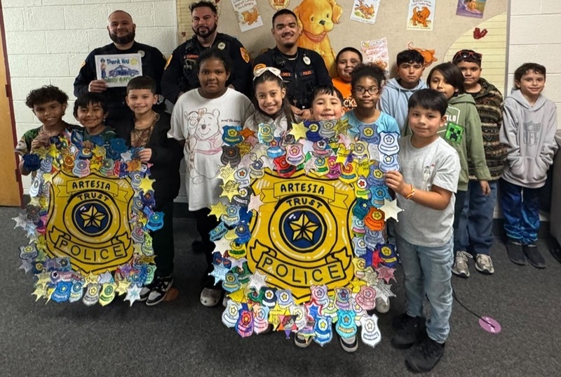 Three male police officers stand behind a group of children who are holding large crafted police badges covered in smaller badges colored by children.