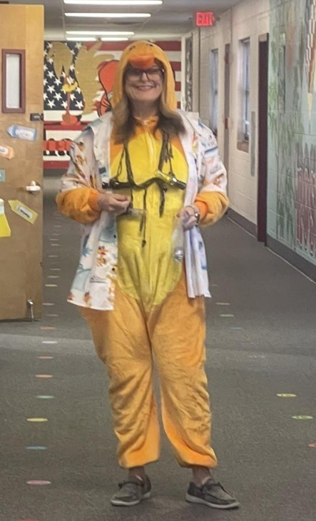 A female teacher poses for a photo in a school hallway. She is dressed as a yellow duck.