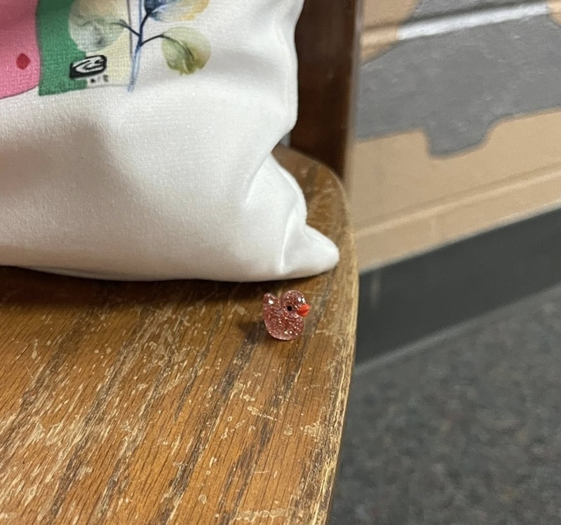 A tiny, pink, glittery plastic duck is pictured sitting on a wooden seat next to a tote bag.