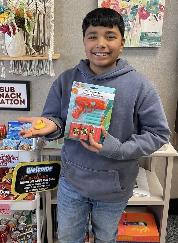 A young boy in a blue-grey hoodie smiles for the camera as he holds a small yellow rubber duck in his palm and a packaged foam dart gun in his other hand.