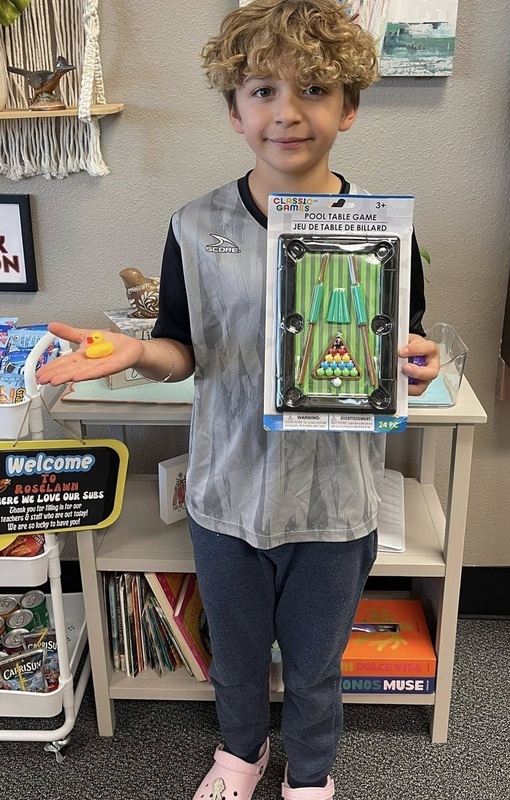 A young boy in a silver and black shirt smiles for the camera as he holds a small yellow rubber duck in his palm and a packaged miniature pool table set in his other hand.