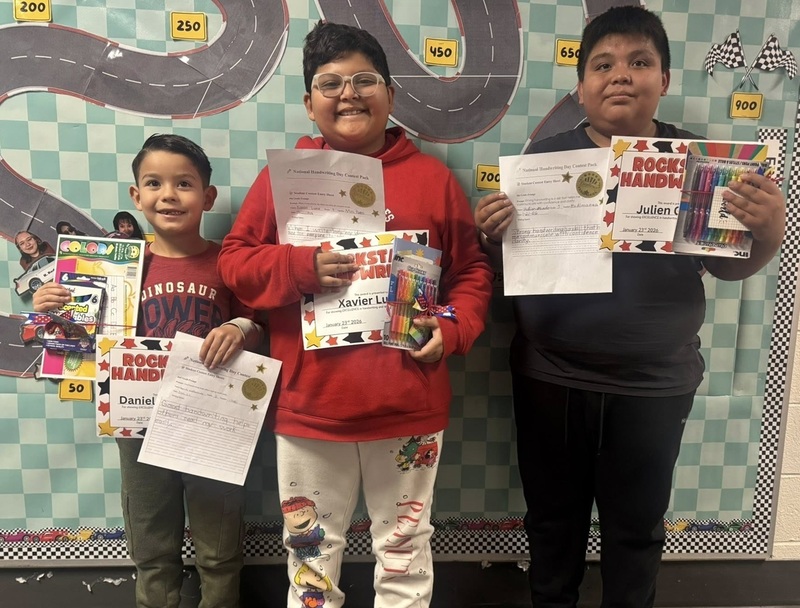 Three young boys smile for the camera and hold up certificates, pens, pencils, and workbooks.