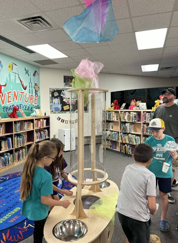 Two young girls stand at a science experiment station featuring a wooden tower and lightweight scarves.