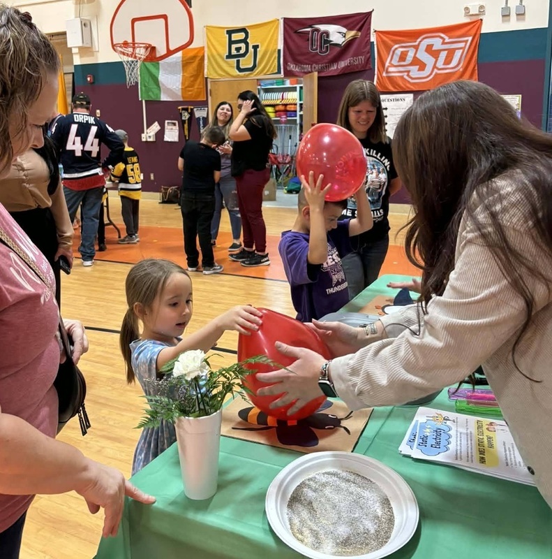 A young girl reaches out to take a red balloon being handed to her by a female teacher at a science experiment station.