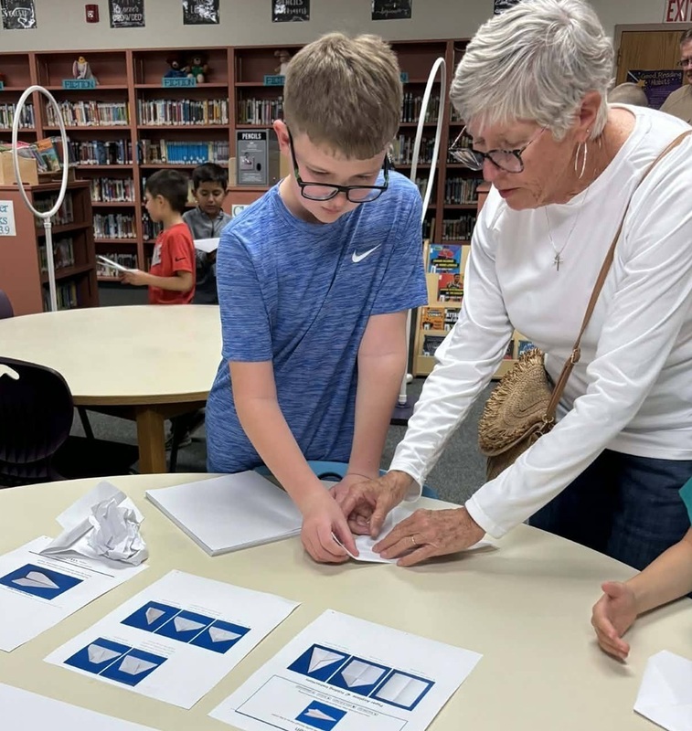A young boy and an elderly woman stand side by side as they work on folding a paper airplane.