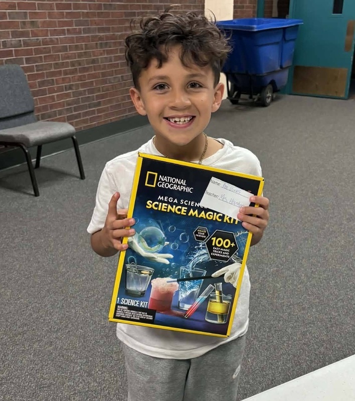 A young boy smiles for the camera as he holds up a science kit.