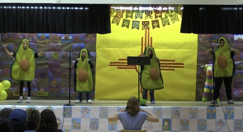 Four young boys dressed as avocados gesture from a stage featuring a large depiction of a New Mexico state flag.