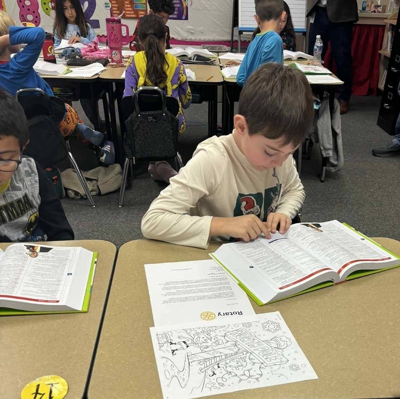 A young boy marks a spot in a book with his finger as he reads while seated at a desk.