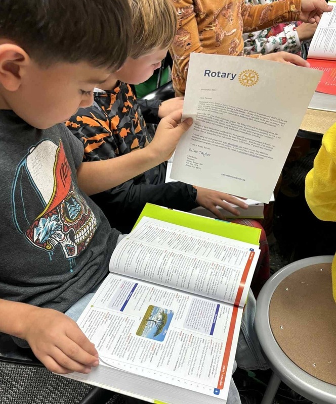 A young boy seated at a desk looks at an open book as he holds a letter in his hand.