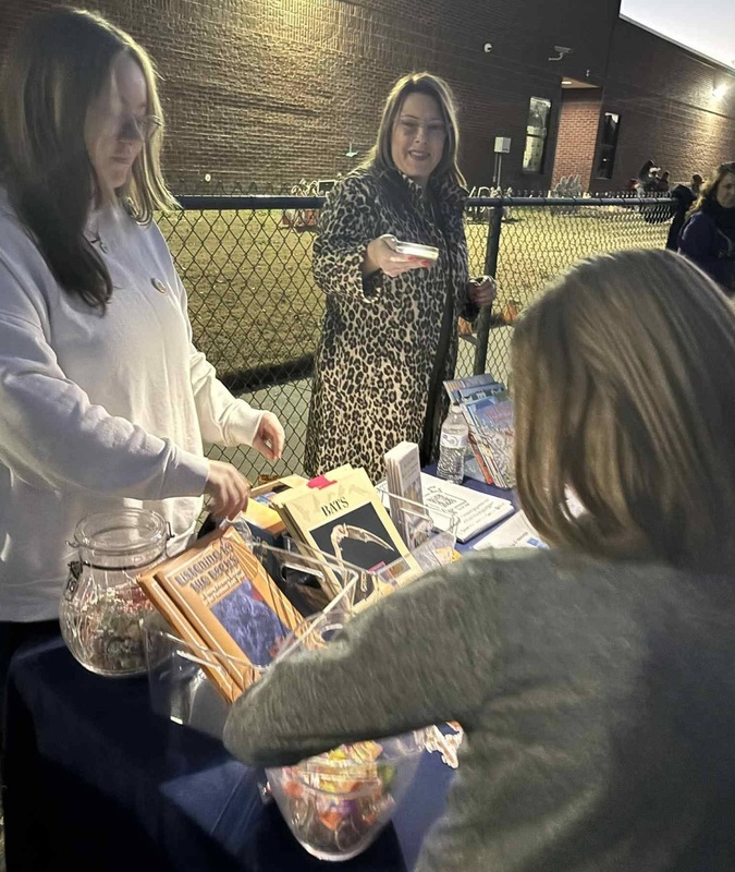 A woman stands behind a table as she speaks to a child about the books placed on it.