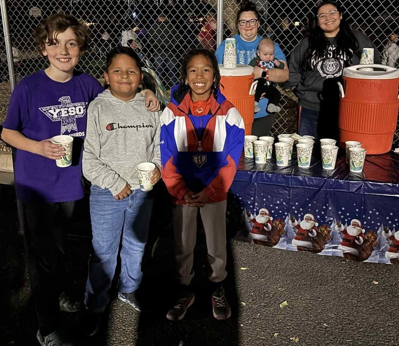Three children pose for a photo in front of a table manned by two women. Large orange water jugs and cups sit on the table.