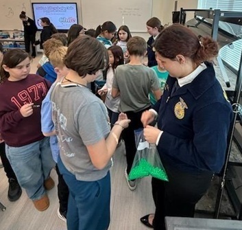A teen girl in an FFA jacket holds a plastic baggie filled with small green items as children in front of her take items from the bag.