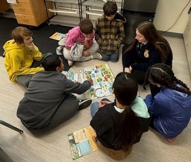 A teen girl in an FFA jacket sits on the floor of a classroom surrounded by young children looking at diagrams.