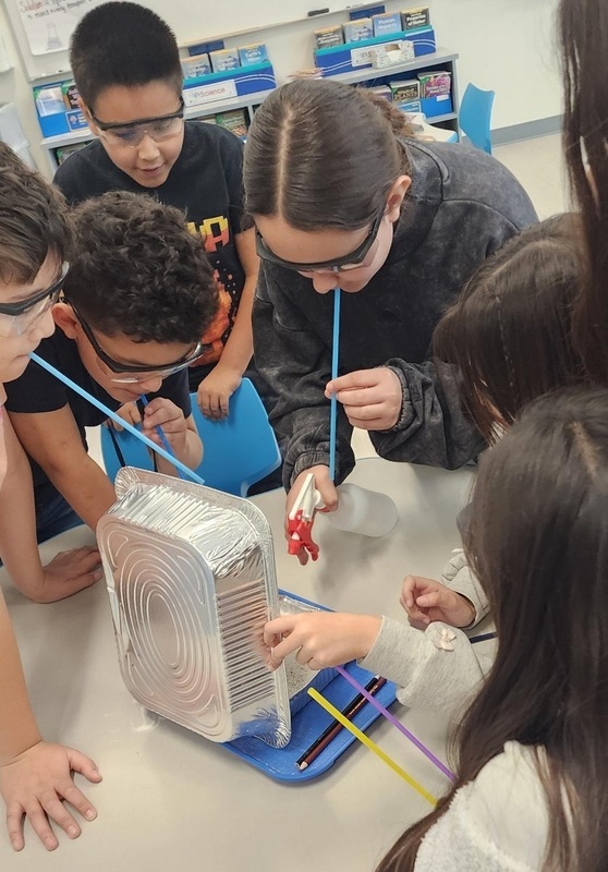 A group of children in goggles use plastic straws to blow on sand in a tin pan.