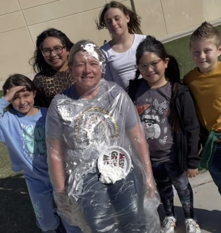One male and four female children pose for a photo around a woman covered in a plastic poncho. The woman has whipped cream in her hair and a pie plate on the plastic poncho.