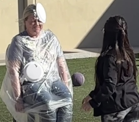 A woman seated on a chair and covered in a plastic poncho cringes as a pie plate thrown by a young girl seen from the back hits her in the head.