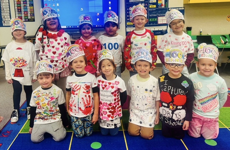 A group of children smile for the camera. They wear "100 Days" hats and shirts decorated with multiples of small items.