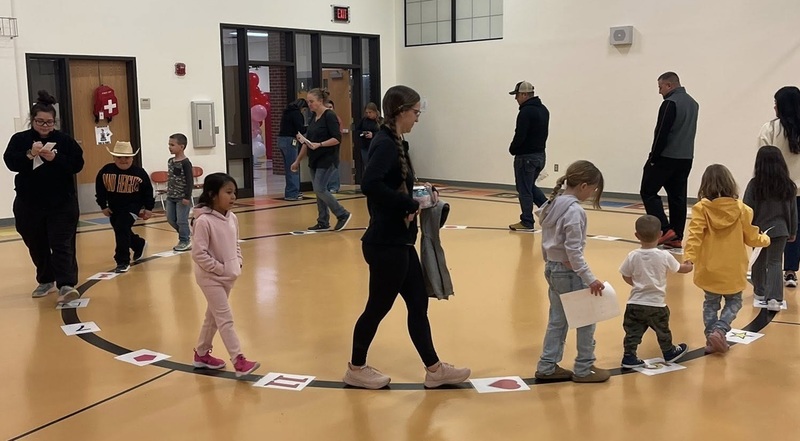 Adults and young children walk around a large numbered circle in a school gym.