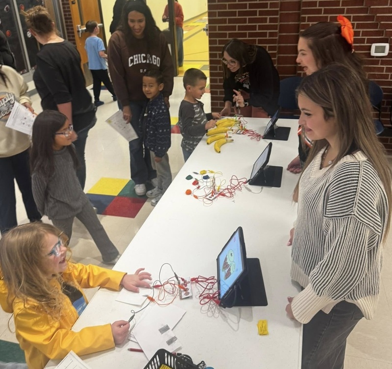 A young girl examines electrodes on a table as a female teacher smiles at her.