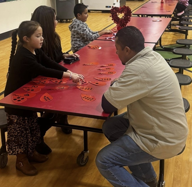A child seated at a long red table moves halves of hearts with black dots on them as a man seated across from her looks on.