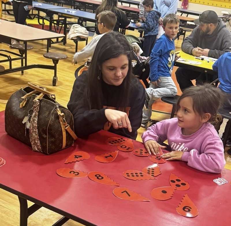 A young girl seated at a long red table with halves of hearts covered in black dots in front of her looks at a woman who is pointing at one of the hearts.