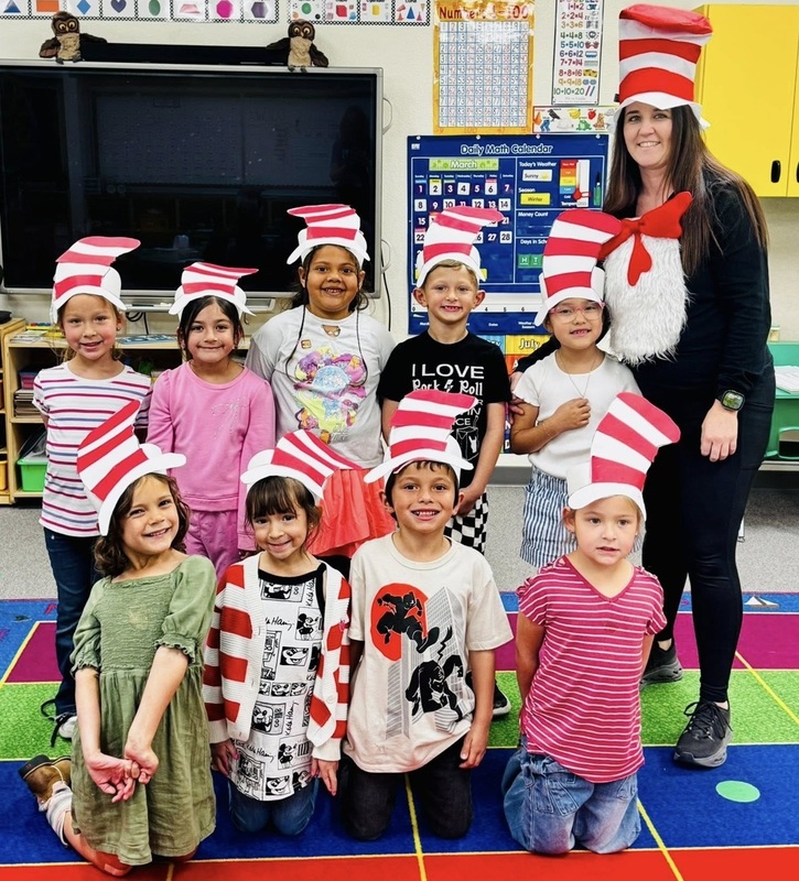 A group of children wearing paper red-and-white-striped hats pose for a photo alongside a female teacher dressed as the Cat in the Hat.