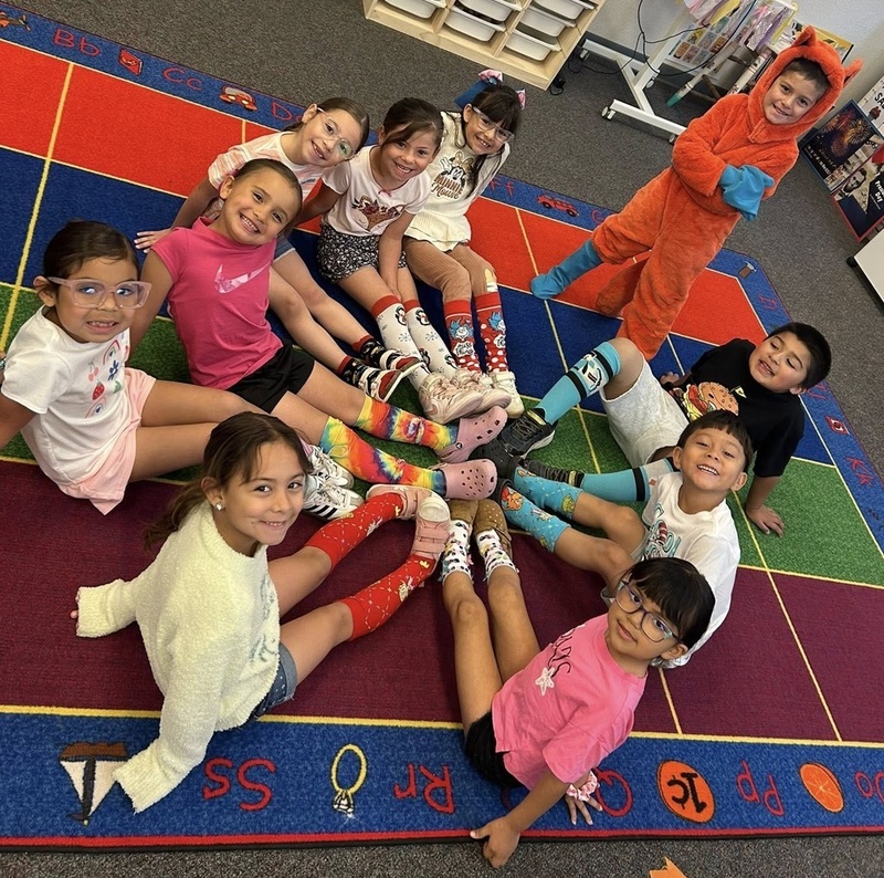 A group of children smiles for the camera as they sit in a circle with their feet together. They are wearing brightly colored socks. A boy dressed as a fox stands beside them.