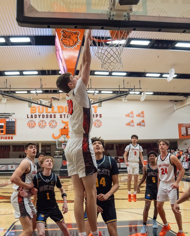A teen boy in a white and orange basketball uniform dunks the ball as three teammates and three opponents in black look on.