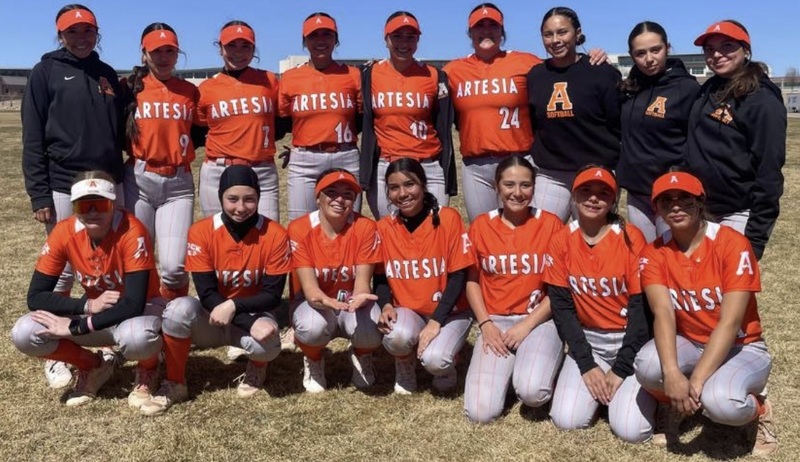 A group of teen girls in orange and grey softball uniforms pose for a photo on a softball field.
