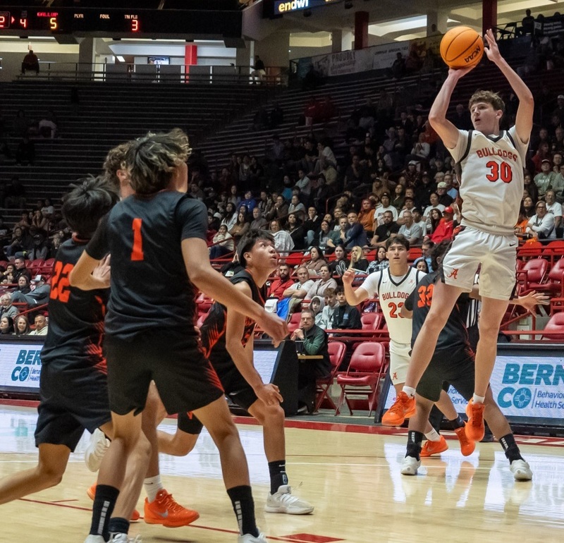 A teen boy in a white and orange basketball uniform holds the basketball over his head in mid-air as he prepares to release it from his right hand. Teammates and opponents in black and orange look on.