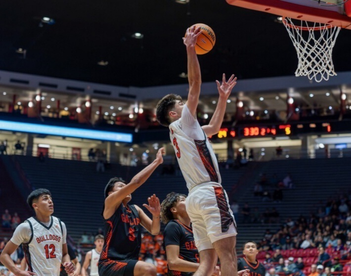 A teen boy in a white and orange basketball uniform goes up in the lane for a layup. An opponent in black and orange is behind him, raising an arm.