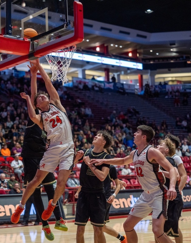 A teen boy in a white and orange basketball uniform leaps while leaning hard to his right side as he lays the ball off the glass ahead of two opponents in black.