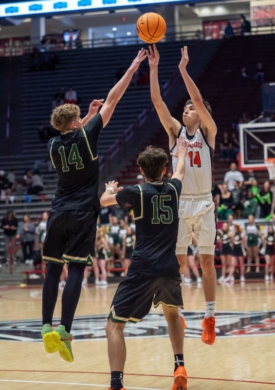 A teen boy in a white and orange basketball uniform jumps and shoots high over a leaping opponent in black whose right arm is extended toward him.
