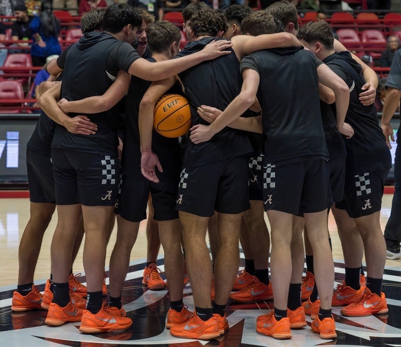 A boys' basketball team in black and orange uniforms huddles at center court with their arms around one another.