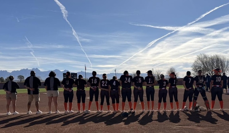 A girls' softball team in black and orange uniforms stands with their backs to the camera along a foul line at a softball field.